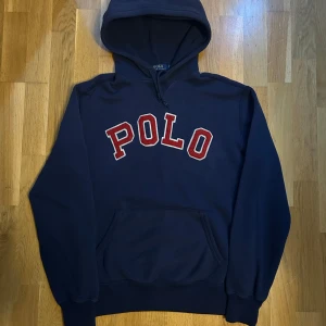 Mörkblå hoodie från Polo Ralph Lauren - Säljer en mörkblå hoodie från Polo Ralph Lauren med stor röd POLO-text framtill. Klassisk känguruficka och huva. 
