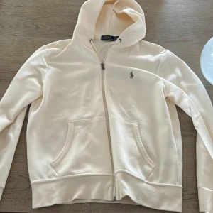 KrämVit hoodie från Polo Ralph Lauren - Säljer en stilren vit hoodie från Polo Ralph Lauren med dragkedja och klassisk broderad logga på bröstet. Tröjan har huva! Köptes in ny för cirka 2000 och den är använd ett fåtal gånger! Passa både s och m