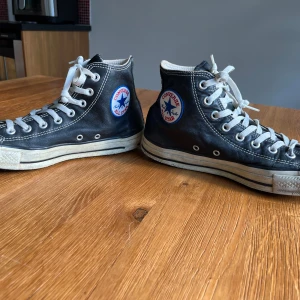 Svarta Converse All Star - Läder svarta Converse All Star high-top sneakers med vit sula och vita snören. Skorna har rund tå, tydliga vita sömmar och den ikoniska loggan på sidan. Perfekta för en avslappnad och tidlös stil. Väl använd , har släppt lite i sidorna