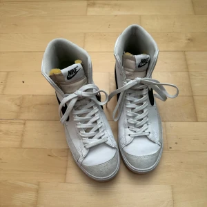 Nike Blazer Mid 77 vita sneakers - Säljer ett par klassiska Nike Blazer Mid 77 i vitt med svart swoosh. Höga sneakers med snörning och retrodesign. Ovandel i skinn och syntet, vit sula och rund tå. Perfekta för dig som gillar streetstyle och tidlös look.