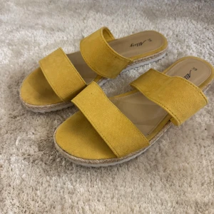 Gula sandaler med dubbla remmar - Säljer ett par trendiga gula sandaler med två breda remmar och platt sula. Skorna har en snygg espadrille-inspirerad kant och är perfekta för varma dagar. Bekväma att glida i och passar till många outfits.