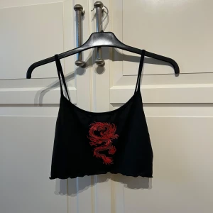 Svart croppad linnetopp med röd drake - Säljer en svart croppad linnetopp med tunna axelband och en stor röd drake tryckt framtill. Toppen är tillverkad i polyester och har en enkel, stilren design med edgy vibe.