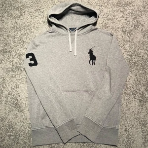 Ralph Lauren hoodie  - Säljer en grå hoodie från Polo Ralph Lauren med svart broderad logga på bröstet och en stor svart siffra 3 på ärmen. Tröjan har huva med vita snören och en klassisk känguruficka framtill. Perfekt för en avslappnad stil.