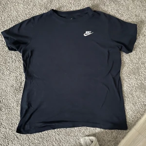 Mörkblå t-shirt från Nike - En klassisk svart t-shirt från Nike med vit logga på bröstet. T-shirten har rund halsringning och korta ärmar. Perfekt för en sportig och avslappnad stil.