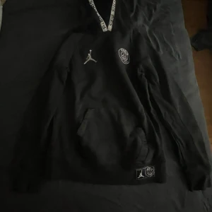 Svart hoodie från Jordan x PSG - Svart hoodie från Jordan och Paris Saint-Germain med vita detaljer och loggor på bröstet och nertill.stl 140-152 har aldrig använts och är som gott som ny