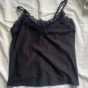 Svart spetslinne med smala axelband - Säljer ett svart linne med smala axelband och vacker spetsdetalj längs urringningen. Toppen har en v-ringad form och är tillverkad i mjukt material, perfekt för lager-på-lager eller som den är. Enkel och stilren design. Ena axelbandet är sönder, kan fixas innan jag postar!
