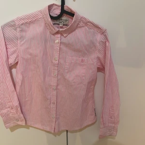 Rosa och vit randig skjorta från Hampton Republic - Säljer en klassisk rosa och vit randig skjorta från Hampton Republic. Skjortan har långa ärmar, bröstficka med broderad logga och knäppning framtill. Perfekt för dig som gillar stilrena och tidlösa plagg.