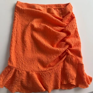 Orange kjol med volang - Säljer en snygg orange kjol med volangkant och rynkade detaljer på sidan. Kjolen har en kort längd och är tillverkad i ett strukturerat syntetmaterial som ger en cool look. Perfekt för dig som vill sticka ut med färg!