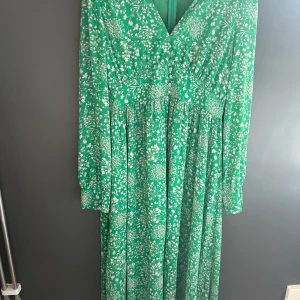 Grön blommig långklänning från Adoore - Säljer en grön långklänning från Adoore med vitt blommigt mönster. Klänningen har v-ringning, långa ärmar och knappar framtill. Perfekt för vår och sommar, med en luftig och feminin känsla.stl 46 men mer som en L/XL. Klänningen är upplagd och går till ankeln, är 165 cm lång 