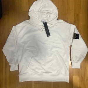 Vit hoodie från Stone Island - Säljer en stilren vit hoodie från Stone Island med klassisk logotyp-patch på vänster ärm. Tröjan har huva med dragsko och en stor magficka framtill. Perfekt för en avslappnad och trendig look.