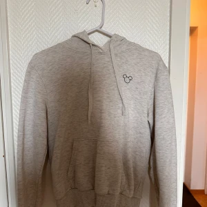 Ljusgrå hoodie med musmotiv från Divided - Ljusgrå hoodie från Divided med en liten svart muskontur broderad på bröstet. Tröjan har huva, dragsko och en stor magficka framtill. Perfekt för en avslappnad stil.