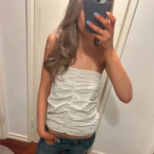 Vit prickig tubtopp - LÅNAD BILD. Säljer en söt vit tubtopp med små svarta prickar och rynkade detaljer framtill. Perfekt till sommaren och enkel att matcha med jeans eller kjol. Toppen är ärmlös och har en tight passform.
