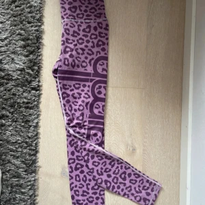 Lila leopardmönstrade leggings aimin  - Säljer ett par lila leggings med leopardmönster och stora tryckta bokstäver längs benet. De är tighta och gjorda i ett stretchigt syntetmaterial, perfekta för träning eller en färgstark vardagslook. Köpta i aimins butik i Halmstad