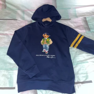 Hoodie från Ralph lauren - Säljer en marinblå hoodie från Polo Ralph Lauren med ett stort tryck av deras ikoniska björn på framsidan. Tröjan har huva, långa ärmar och två gula ränder på ena ärmen. Perfekt för dig som gillar streetwear och klassiska märken.