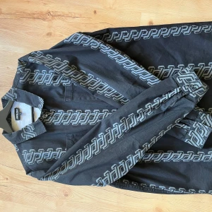 Svart skjorta med grått mönster från Santa Cruz - Säljer en svart överskjorta/jacka från Santa Cruz med unikt grått grafiskt mönster över hela plagget. Skjortan har klassisk krage, dragkedja framtill och långa ärmar. 