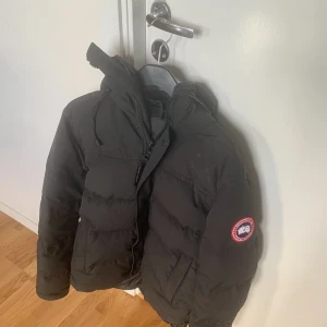 Svart dunjacka från Canada Goose - Säljer en svart dunjacka från Canada Goose med huva och klassisk logotyp på ärmen. Jackan är tjock, har dragkedja framtill och är perfekt för kalla dagar. Snygg och stilren design som håller dig varm hela vintern. McMillan parka modellen 