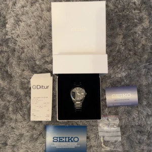 Seiko herrklocka med svart urtavla och stållänk - Stilren Seiko klocka med svart urtavla, datumvisning och silverfärgad stållänk. Klockan levereras i originalask med extra länkbitar och manual. Perfekt för dig som gillar klassisk design och kvalitet.