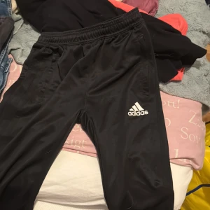 Svarta träningsbyxor från Adidas - Svarta träningsbyxor från Adidas med klassisk logga på låret och elastisk midja. Byxorna har raka ben och är gjorda i ett glansigt syntetmaterial som passar perfekt till sport och träning.