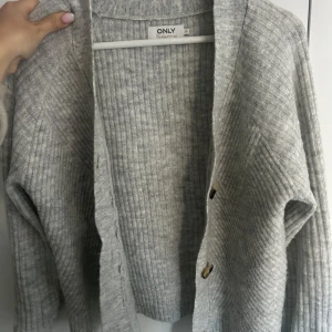 Ljusgrå ribbad kofta från ONLY - Mysig ljusgrå kofta från ONLY med ribbad struktur och två knappar framtill. Perfekt att slänga på sig över en t-shirt eller linne. Långärmad och i mjukt material.