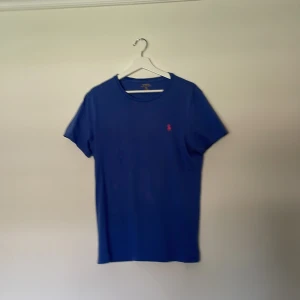 Blå t-shirt från Polo Ralph Lauren - Säljer en blå t-shirt från Polo Ralph Lauren i modellen Custom Fit. T-shirten har rund halsringning och en liten röd broderad logga på bröstet. Perfekt för en stilren och klassisk look.