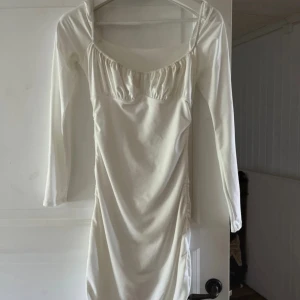 Vit långärmad klänning med rynkade detaljer - Säljer en stilren vit klänning med lång ärm och fyrkantig ringning. Klänningen har rynkade detaljer vid bysten och längs sidorna vilket ger en snygg passform. Perfekt för dig som gillar en enkel men trendig look. Är köpt i storlek S men passar även en som är i Xs, använd 1 gång