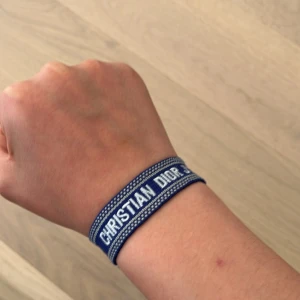 Blått armband från Christian Dior - Säljer ett blått tygarmband från Christian Dior med texten 'CHRISTIAN DIOR J'ADIOR' i vitt. Armbandet har dekorativa tofsar i ändarna och ett vävt mönster längs kanterna. Perfekt accessoar för att lyfta din stil.