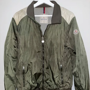 Grön bomberjacka från Moncler - Säljer en grön bomberjacka från Moncler. Den har en dragkedja framtill och ribbade muddar vid ärmslut och krage. Jackan är i acceptabelt skick och har några små skador på ena ärmen. Perfekt för höst och vår!