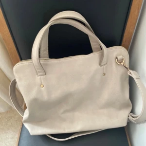 Axelväska med dragkedja - Snygg beige axelväska med både handtag och justerbar axelrem. Väskan har guldfärgade detaljer och stängs med dragkedja. Insidan är rymlig med en innerficka. Perfekt för att bära med sig det viktigaste i vardagen.