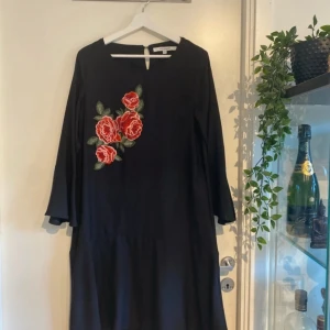 Svart klänning med broderade rosor - Svart klänning med trekvartsärm och vackra broderade röda rosor med gröna blad på framsidan. Klänningen har en rak passform och rund halsringning. Perfekt för dig som vill sticka ut med en unik detalj.