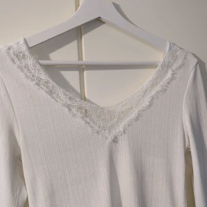 Vit ribbad topp med spets från H&M - Säljer en vit långärmad topp från H&M i storlek S. Toppen har ribbat material och v-ringning med fina spetsdetaljer längs halsen. Perfekt att styla med jeans eller kjol för en fräsch look.