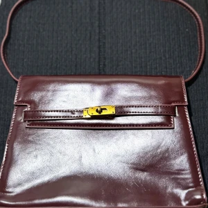 Vinröd axelväska i skinn med gulddetaljer - Säljer en stilren vinröd axelväska i skinn med elegant guldfärgat spänne framtill. Väskan har en klassisk rektangulär form och ett rymligt huvudfack. Perfekt för dig som vill ha en tidlös accessoar med lyxig känsla. Helt oanvänd!