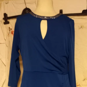 Blå klänning med glittrig krage från Happy Holly - Snygg blå klänning från Happy Holly med lång ärm och glittrig detalj runt halsen. Klänningen har en liten öppning fram och markerad midja för en smickrande siluett. Perfekt för dig som vill sticka ut lite extra. Storlek 40/42