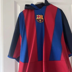 FC Barcelona fotbollströja Ronaldinho #10 - Klassisk FC Barcelona fotbollströja med rödblåa vertikala ränder, klubbmärke på bröstet och Ronaldinho 10 i gult på ryggen. Tröjan har huva och korta ärmar, perfekt för fans av laget och legenden. Retro 