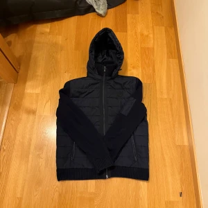 Cardigen jacka pier one - Säljer en svart pufferjacka med huva och dragkedja framtill. Jackan har quiltad design, ribbade muddar och en extra ficka med dragkedja på ärmen. Perfekt för kyligare dagar.