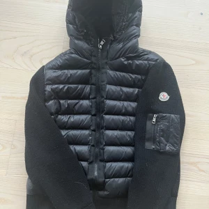 Svart cardigan med huva från Moncler - Säljer en svart Moncler-jacka med quiltad front och stickade ärmar. Jackan har huva, dragkedja och en ficka med dragkedja på ena ärmen. Perfekt för lager-på-lager och snygg streetstil. Skriv gärna vid eventuella frågor eller för mer bilder. Pris kan diskuteras vid snabb affär!
