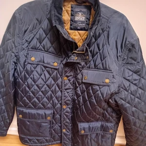 Svart quiltad jacka från Hampton Republic - Säljer en svart quiltad jacka från Hampton Republic med guldfärgade knappar och fyra fickor framtill. Jackan har lång ärm och ståkrage, samt ett brunt innerfoder. Perfekt för kyliga dagar.