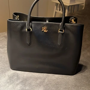 Svart handväska från Ralph Lauren - Snygg svart handväska från Ralph Lauren med guldfärgade detaljer och RL-logga framtill. Väskan har två handtag, avtagbar axelrem och dragkedja i mittenfacket. Perfekt för att bära dina viktigaste saker med stil.