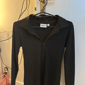 Svart ribbad långärmad  från Kappahl - Svart långärmad polotopp från Kappahl med ribbad struktur och djup v-ringning. Klassisk krage och knappar framtill. Perfekt för lager-på-lager eller som den är.