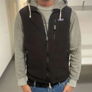 Svart fleeceväst från Patagonia - Svart fleeceväst från Patagonia med dragkedja framtill och tre fickor med dragkedja. Klassisk modell med hög krage och mjukt material. Perfekt att ha över en hoodie eller tröja.