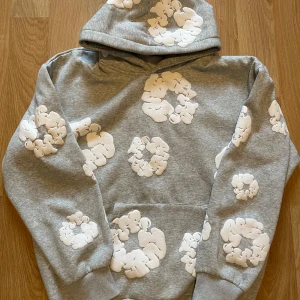 Grå hoodie med vita blommor från Denim Tears - Säljer en grå hoodie från Denim Tears med vita blommönster över hela tröjan. Hoodien har en klassisk passform, huva och magficka. Perfekt för dig som vill sticka ut med en unik streetwear-stil.