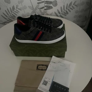Gucci Ace Sneakers  - Säljer ett par snygga Gucci sneakers i svart med klassiskt monogrammönster och röd/blå rand på sidan. Skorna har vit platt sula, svarta skosnören och detaljer i rött och blått på hälen. Perfekta för dig som vill sticka ut med stil. OBS! PRISET KAN DISKUTERAS VID EN SNABB AFFÄR!