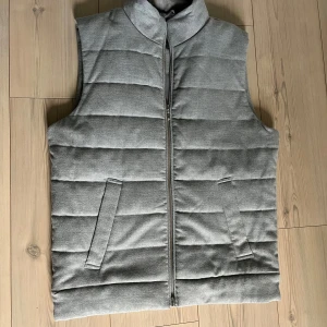 Grå quiltad dunväst  - Snygg grå dunväst med quiltad design och hög krage. Västen har två snedställda fickor framtill och stängs med dragkedja. Perfekt för lager-på-lager och enkel att matcha med olika outfits. Den är från dressman och är sällsynt att hitta, liknar em stenströms väst. Väldigt fräsch överlag och perfekt nu till våren. 