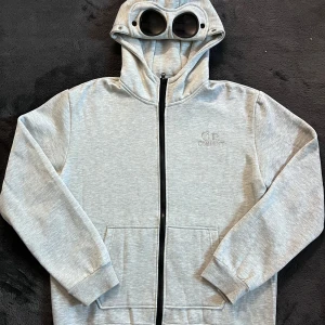 C.P. Company zip Hoodie - Säljer en helt ny ljusgrå hoodie från C.P. Company med unika inbyggda glasögon i huvan. Tröjan har dragkedja framtill, två fickor och ribbade muddar. Skick 10/10 helt ny, storlek L, kan ej garantera att den är äkta då den e köpt på plick och saknar qr kod. Pris går att diskutera✅
