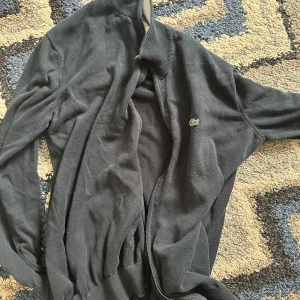 Svart cardigan jacka från Lacoste - Snygg svart cardigan jacka från Lacoste med dragkedja framtill och ribbade muddar. Klassisk design med lång ärm och diskret logga på insidan av kragen. Perfekt för en stilren look.