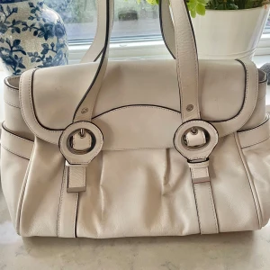 Vit handväska från Nine West - Säljer en stilren vit handväska från Nine West med stora runda spännen och långa handtag. Lite 60-70 tal känsla! 💗Väskan har ett randigt innerfoder i brunt, blått och gult samt en innerficka med dragkedja. Väskan är i bra skick fast använd!💗