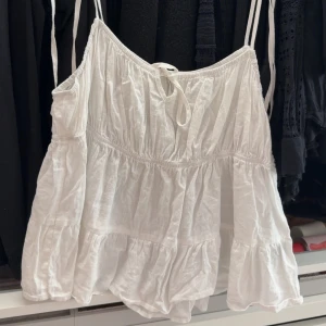 Vit volangig linnetopp med knytband - Söt vit linnetopp med tunna axelband och volangdetaljer. Toppen har knyt framtill och är luftig med en avslappnad passform, perfekt för varma dagar.