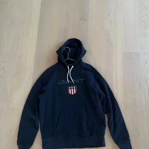Mörkblå hoodie från GANT - Säljer en mörkblå hoodie från GANT med vit dragsko och broderad logga samt sköld på bröstet. Klassisk känguruficka framtill och mjukt material, perfekt för en avslappnad stil.