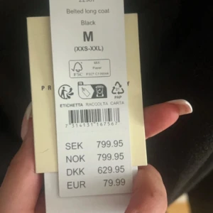 Svart lång kappa med bälte - Säljer en svart lång kappa med bälte i midjan. Klassisk krage och enkel design som ger en stilren look. Perfekt för dig som vill ha en tidlös jacka med snygg passform.