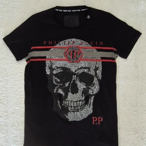 Philipp plein Svart t-shirt S - Cool svart t-shirt från Philipp Plein med glittrande döskallemotiv framtill och röda detaljer. T-shirten har rund hals och korta ärmar. Märkets logga syns både på bröstet och i nacken. I Mycket fint skick, storlek S