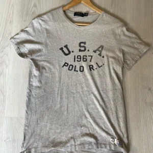 Grå t-shirt från Polo Ralph Lauren - Säljer en grå t-shirt från Polo Ralph Lauren med tryck 'U.S.A. 1967 POLO R.L.' på bröstet. Klassisk rund halsringning och korta ärmar. Perfekt för en avslappnad stil🔥. Se defekter bild 4/5